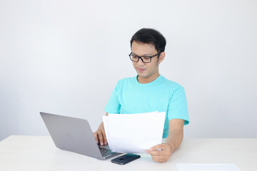 Young Asian Man is smile and happy when working on a laptop and document on hand. Indonesian man wearing blue shirt.