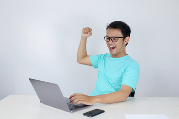 Happy excited and smiling young Asian man raising his arm up to celebrate success or achievement when working in laptop. Indonesian man wearing blue shirt.