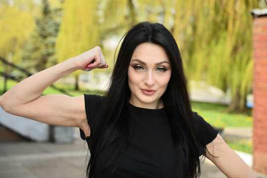Fitness model posing against the backdrop of a city park in fine weather. Photo in spring weather outdoors, a brunette girl with a slender figure shows biceps arms.