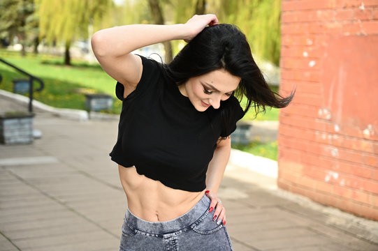 Photo in spring weather outdoors, a caucasian brunette girl with a slim figure is posing with pleasure. Fitness model stands against the backdrop of a city park in fine weather.