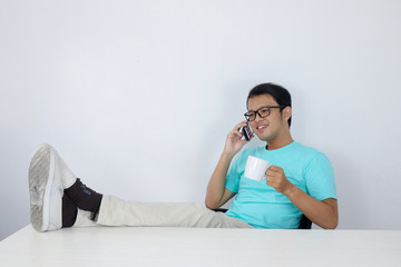 Fototapeta na wymiar Young Asian man is feeling happy and smile face when talk on the phone with leg on the table.