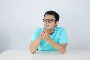 Young Asian man wear blue shirt and glasses with serious face looking on the camera and hand hold chin on the table.