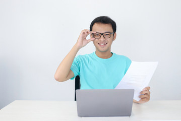 Young Asian Man is smile and happy when working on a laptop and document. Indonesian man wearing blue shirt.