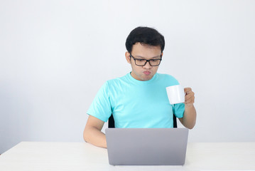 Young Asian man is serious when work in laptop with drink coffee in white glass. Indonesia Man wear blue shirt Isolated grey background.