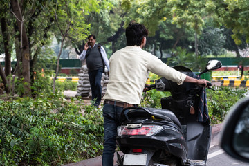 A Indian man pushing his bike as he walks along due to scooter breakdown on the streets. Rear view...