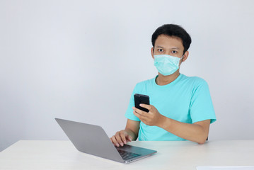 Fototapeta na wymiar Young Asian Man wearing medical mask is serious and focus when working on a phone and laptop on the table. Indonesian man wearing blue shirt.