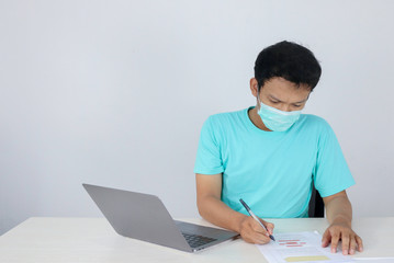 Young Asian man wearing medical mask is feeling unhealthy, tired, and confused with working in documents and laptops on the table.