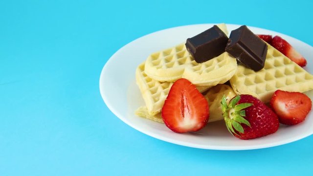 Waffles on plate with two chocolate cubes and strawberries on blue background