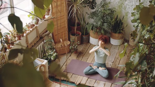 High angle shot of woman sitting in lotus pose on exercise mat, holding hands in prayer gesture and then in mudras while having yoga meditation in cozy room with wooden floor and green plants