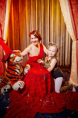Obraz na płótnie Canvas Family during a stylized theatrical circus photoshoot in a beautiful red location. Models mother who looks like a trainer and daughter who looks like a tiger pose on stage with curtains