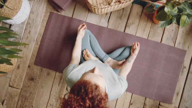 Top view zoom out shot of barefoot woman with red curly hair sitting in lotus pose on mat and holding hands in mudras while practicing yoga meditation at home in room with wooden floor