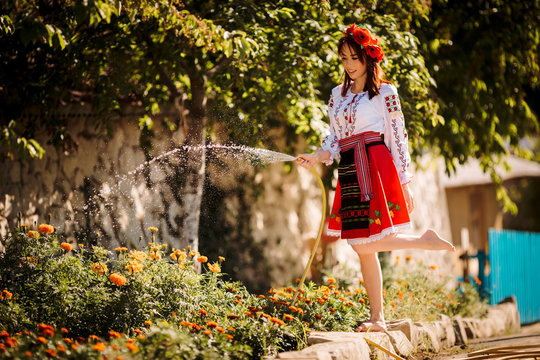 Young redhead moldavian girl dressed in traditional costume with flower wreath on the head watering  flowers