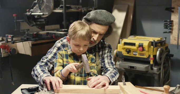 Caucasian grandfather carpenter teaching small cute grandson to spike with hammer in workshop. Old woodman showing little boy how to drive a nail and work with wood and timbers.