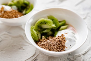 A pair of small dessert bowls with fresh kiwi fruit, white ice cream, yogurt and chocolate chips on a white stone background with teaspoons