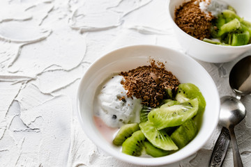A pair of small dessert bowls with fresh kiwi fruit, white ice cream, yogurt and chocolate chips on a white stone background with teaspoons