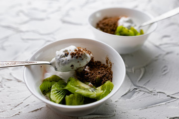 A pair of small dessert bowls with fresh kiwi fruit, white ice cream, yogurt and chocolate chips on a white stone background with teaspoons