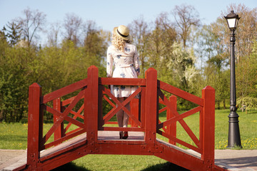 Beautiful woman in a white summer dress and a straw hat stands on a birch bridge in the park