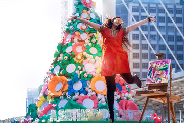 Street female artists feel happy, jumping and hand up in a modern city, big Christmas tree in background