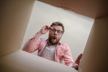 Surprised man looks into the unpacked delivered box with the order, hold glasses with hands and rejoices. Unboxing inside view. - obrazy, fototapety, plakaty