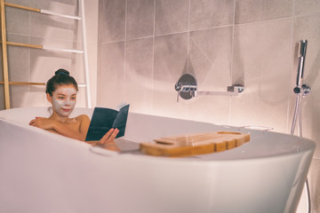 Woman taking a hot bath in modern bathtub bathroom relaxing reading a book while doing a clay face...