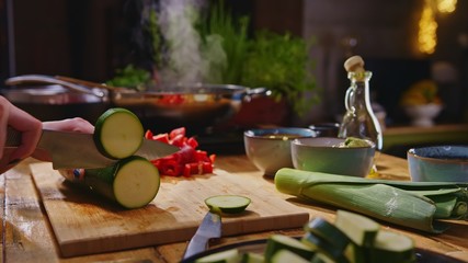 Woman slicing courgette and vegetables for cooking on kitchen table. Closeup hands. Cosy dark room. Real, authentic cooking.