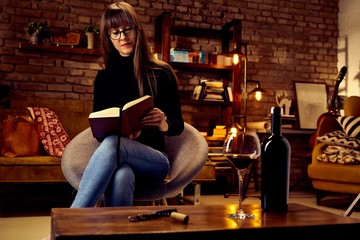 young woman drinking wine at loft home while reading a book.