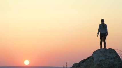 Silhouette of a woman hiker jumping alone on big stone at sunset in mountains. Female tourist raising her hands up on high rock in evening nature. Tourism, traveling and healthy lifestyle concept.