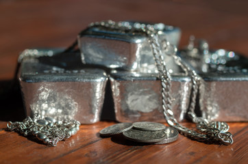 A pile of various silver coins, silver bars and jewelry on a mahogany table. Selective focus.