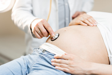 Doctor listening to a pregnant woman's belly with a stethoscope during a medical examination, close-up view