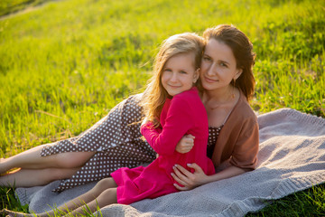 Beautiful young mother and her little daughter in pink dress having fun on nature. They are sitting on a plaid, hugging and smiling. Maternal care and love. Horizontal photo