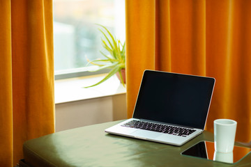 A laptop computer laying on a green couch and a orange curtain. A retro interior with warm colors and a cup of tea on the side. Modern technology and vintage retro mix