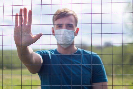 Handsome guy in protective mask on face standing behind bars, lattice. Restriction of freedom. Self isolation, coronavirus epidemic virus infection. Covid-19. Isolated young man show palm, stop sign