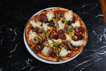 Pizza with arugula decoration consisting of sausage, mushrooms, corn, sausage, black olives, tomatoes and cheddar cheese slices on a black marble table