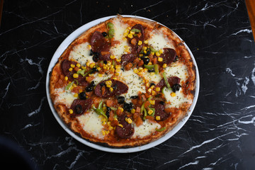 Pizza with arugula decoration consisting of sausage, mushrooms, corn, sausage, black olives, tomatoes and cheddar cheese slices on a black marble table