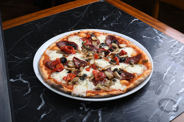 Pizza with arugula decoration consisting of sausage, mushrooms, corn, sausage, black olives, tomatoes and cheddar cheese slices on a black marble table