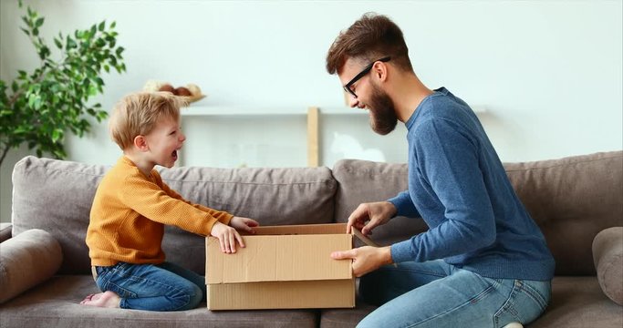 Side view of cheerful boy and bearded man opening cardboard box and laughing while sitting on couch at home