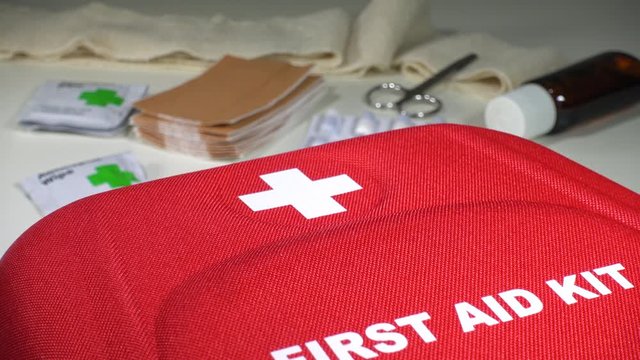 Close POV reverse dolly shot of a first aid kit on a clean surface, with a red first aid box, sticking plasters, bandage, antiseptic wipes, scissors and medication.