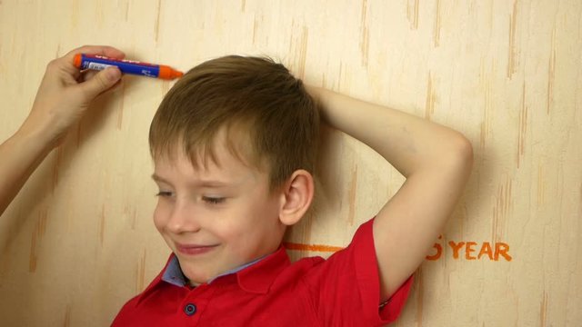 A cute Caucasian boy stands near a wall on which growth marks are 5 years old, mom’s hand draws a new growth mark. Fast growing kids