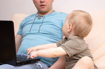 Naklejka premium Little baby sitting on father's knees at his workplace. Man holding child in front of laptop and working remotely.