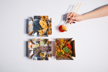 top view of woman holding chopsticks with dumpling near sauce and takeaway boxes with prepared chinese food on white