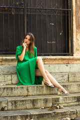 Fototapeta na wymiar Portrait of a sexy Caucasian woman in a casual green dress sitting on stairs in old town. Elegant girl resting in the steps of a church. Concept of travel, holidays, fashion and style.