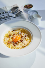 delicious pasta carbonara served with cutlery, cream and seasoning on white table in sunlight