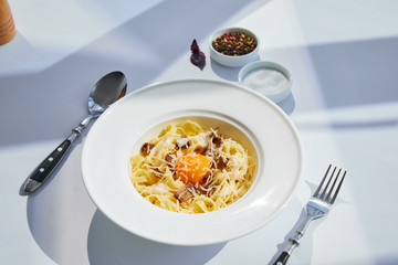 delicious pasta carbonara served with cutlery and seasoning on white table in sunlight