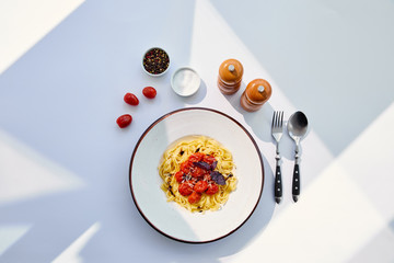 top view of delicious pasta with tomatoes served with cutlery, salt and pepper mills on white table in sunlight