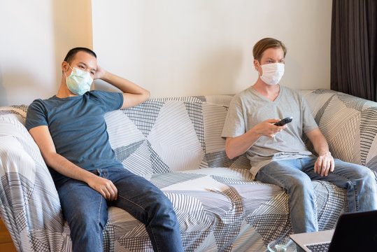 Two multi ethnic men with mask as friends watching tv in quarantine at home