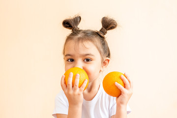 Fototapeta na wymiar A little girl in a white T-shirt holds two orange oranges in her hands. A girl in a white T-shirt laughs and holds two oranges in her hands. A child in a white T-shirt holds two oranges on a beige bac