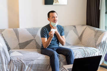 Stressed young Asian man with mask having sore throat at home under quarantine