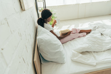 high angle view of african american girl sitting in bed and reading book