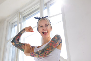 Sunny shot of joyful young pretty blonde woman with tattoos showing cherfully tongue to camera while demonstrating her strong biceps, posing in front of big window