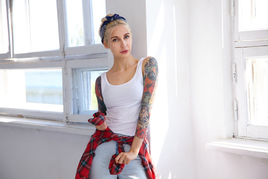 Indoor shot of young lovely tattooed blonde lady dressed in casual clothes while standing in front of big window on bright sunny day and looking attentively at camera